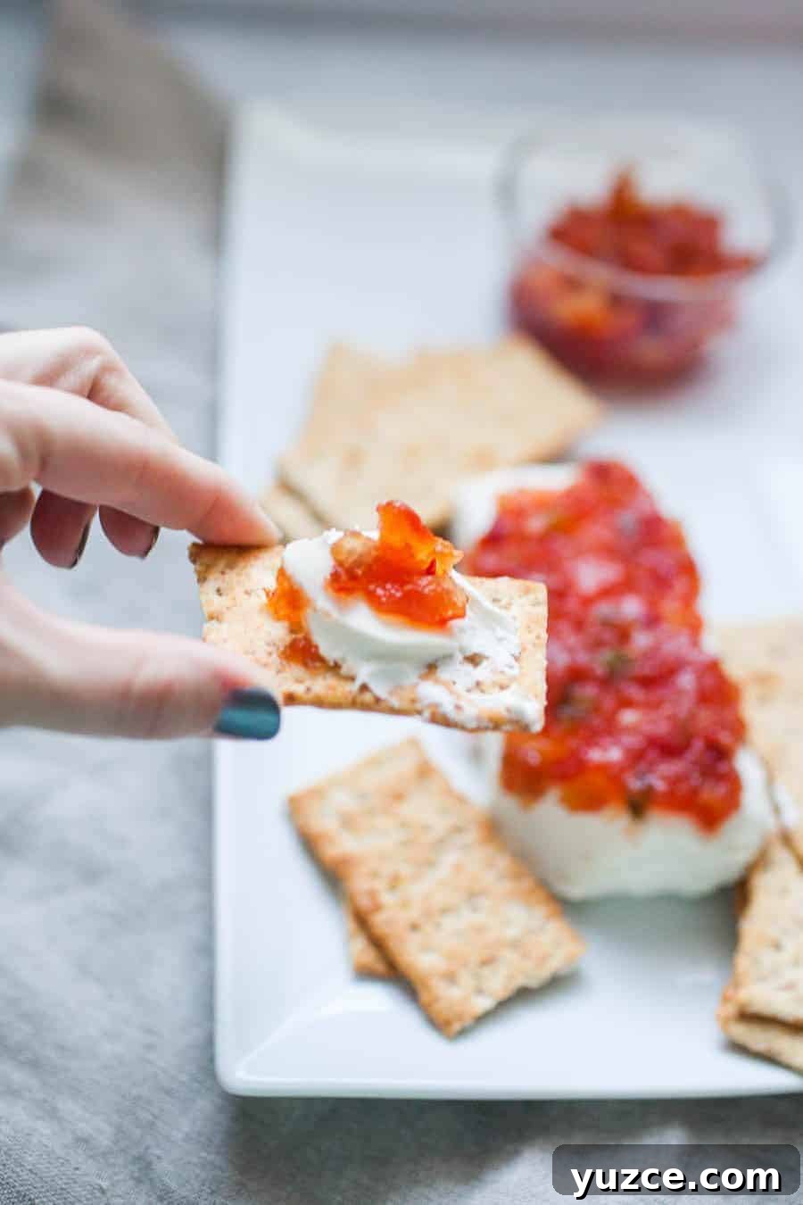 Sweet Pepper Jelly and Cream Cheese Dip Recipe for Entertaining 2 A hand holding a cracker topped with cream cheese and red pepper jelly above a platter with more crackers, cream cheese, and pepper jelly.