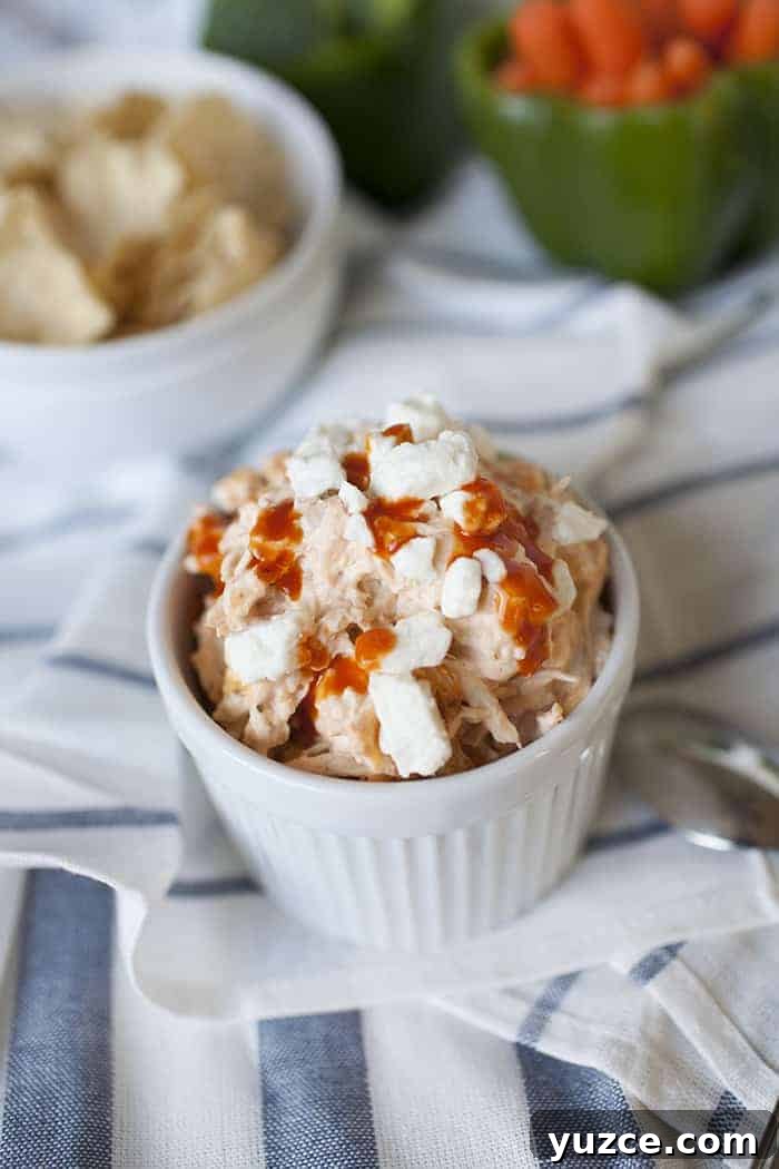 Sweet Pepper Jelly and Cream Cheese Dip Recipe for Entertaining 11 A small white bowl of buffalo chicken dip topped with crumbled cheese and hot sauce, placed on a striped cloth next to a bowl of chips and a green bell pepper filled with vegetables.