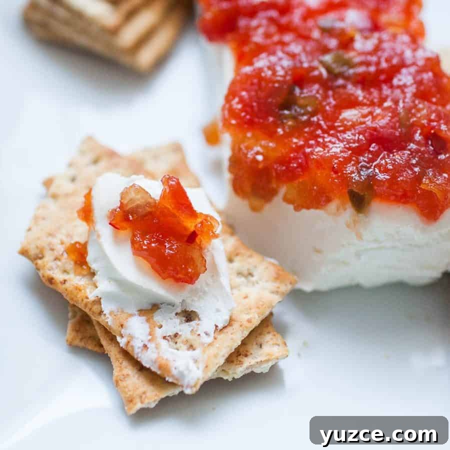 A close-up of crackers topped with cream cheese and a reddish pepper jelly, alongside a block of cheese with the same spread on a white plate.