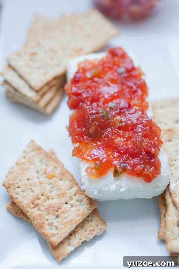 Sweet Pepper Jelly and Cream Cheese Dip Recipe for Entertaining 5 A block of pepper jelly and cream cheese is served on a white plate with a side of square crackers.