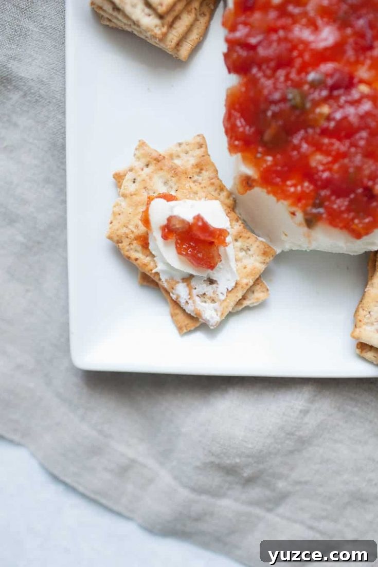 Sweet Pepper Jelly and Cream Cheese Dip Recipe for Entertaining 6 A plate with a cheese ball covered in red salsa, accompanied by a stack of crackers. One cracker is topped with pepper jelly and cream cheese. The setup is on a gray cloth.