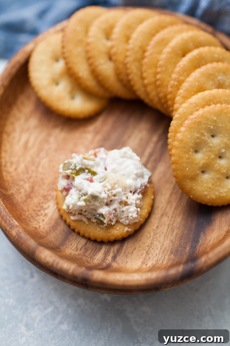 Sweet Pepper Jelly and Cream Cheese Dip Recipe for Entertaining 9 A brown wooden plate holding a row of round crackers, with one cracker topped with a mixture of creamy white spread, chopped vegetables, and jalapeño popper dip.