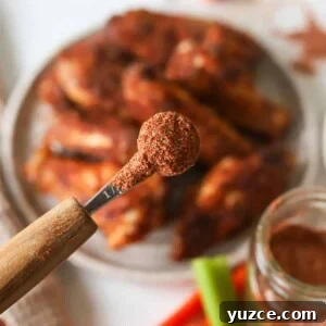 A close up shot of buffalo chicken dry rub in a measuring spoon above a plate of buffalo chicken wings.