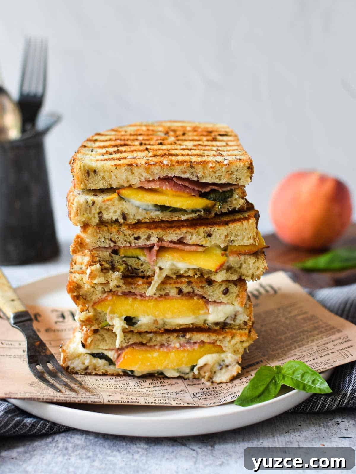 Peach, Prosciutto & Burrata Panini Recipe for Summer Sandwiches 7 Eye level view of peach, prosciutto and burrata panini stacked high on a white plate with a peach in the background.