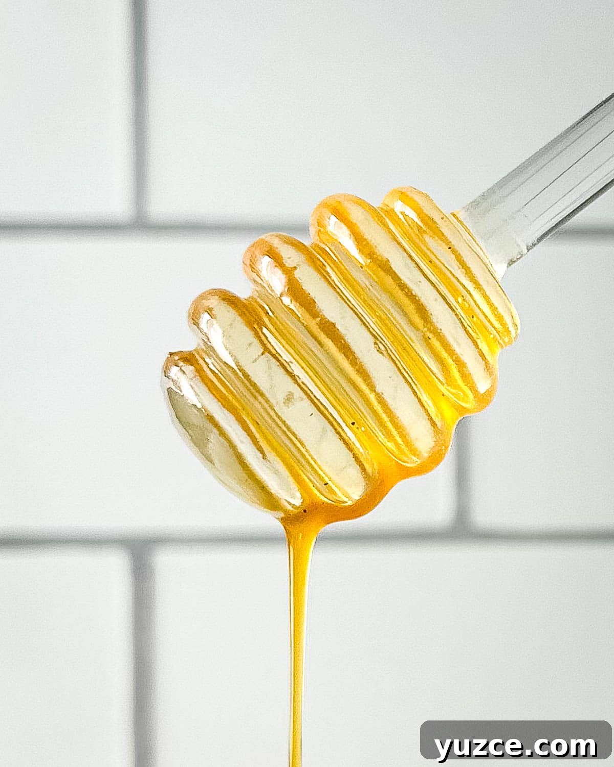 Homemade Hot Honey Sauce Recipe for Spicy-Sweet Dishes 2 Closeup of honey dipper dripping hot honey sauce in front of a white subway tile wall.
