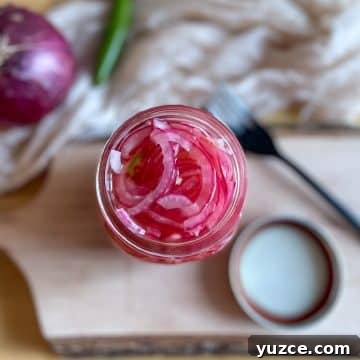 Sliced red spicy pickled onions are shown in a glass jar on a rustic wood cutting board with a lid, a black fork, a white linen, a red onion, and a Serrano pepper in the background.