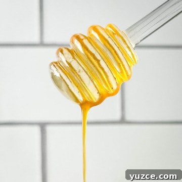 Homemade Hot Honey Sauce Recipe for Spicy-Sweet Dishes 13 Closeup of honey dipper dripping hot honey sauce in front of a white subway tile wall.