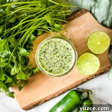 A jar of honey jalapeno lime vinaigrette sits on a cutting board surrounded by limes, cilantro, and jalapenos.
