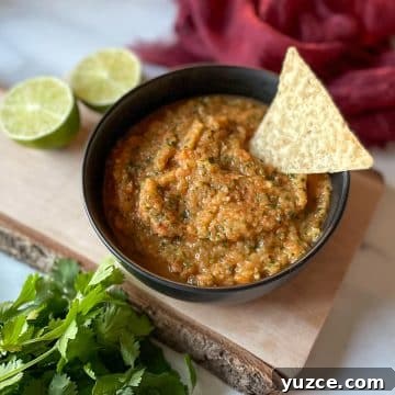A bowl of pineapple habanero salsa sits on a rustic wooden cutting board surrounded by limes, cilantro, and a red linen.