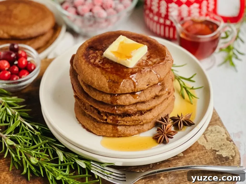 Gingerbread Pancakes with Warm Molasses Syrup 4 A white plate filled with dark brown colored round pancakes with a large square of melty butter and syrup running down the top and a small bowl of cranberries and syrup nearby.