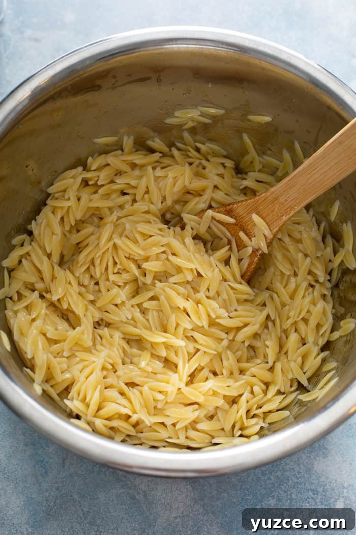 Greek Orzo Salad with Feta, Cucumber & Lemon Dressing 5 Cooked orzo pasta being stirred with a wooden spoon in a metal mixing bowl