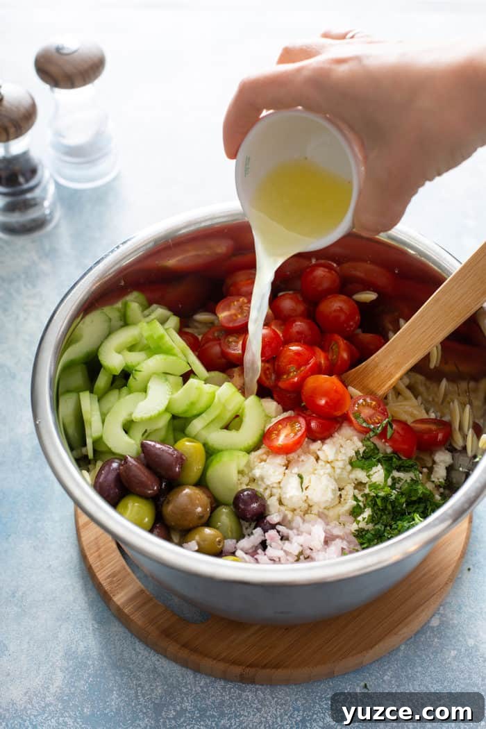 Greek Orzo Salad with Feta, Cucumber & Lemon Dressing 7 Dressing being poured over greek orzo salad in a metal mixing bowl