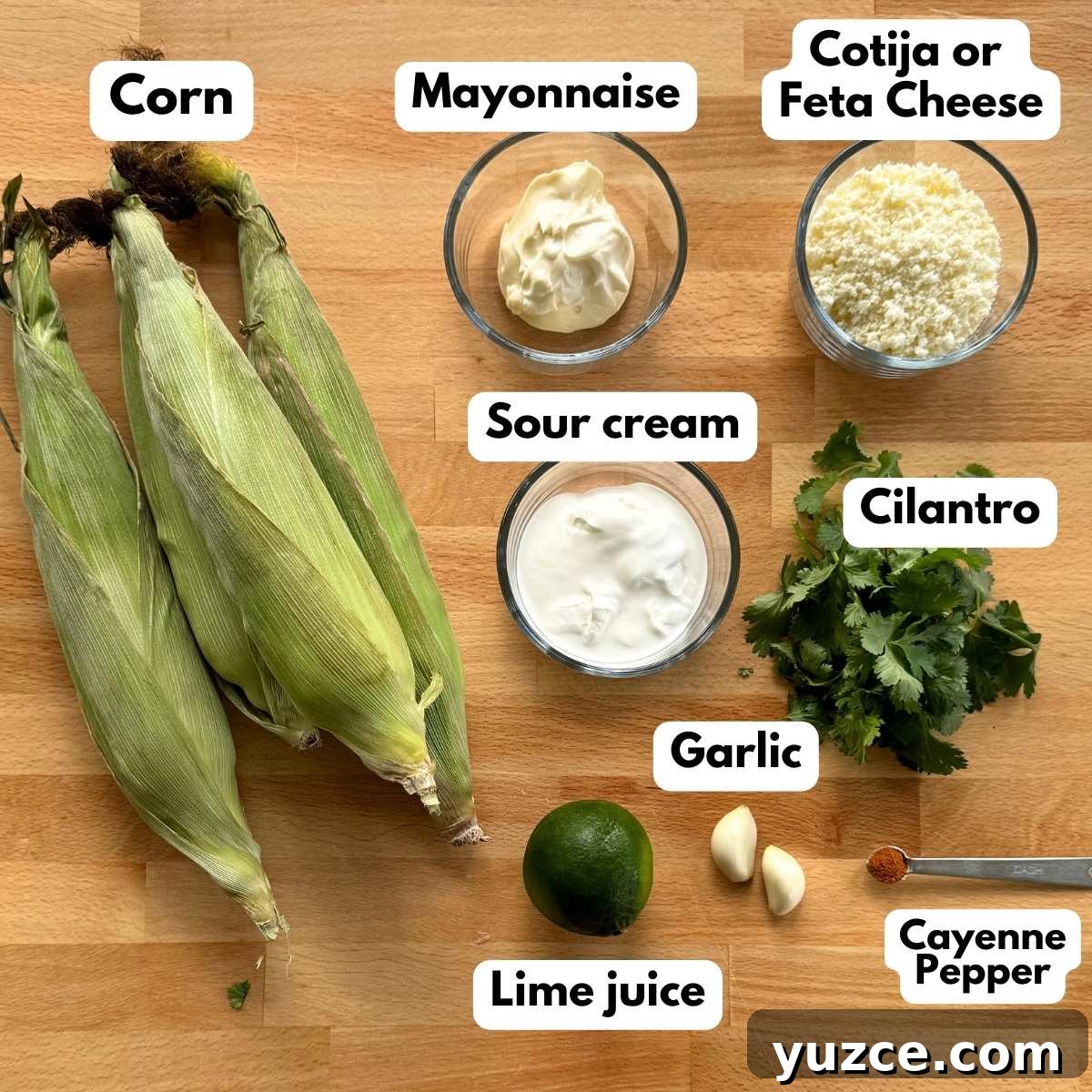 Labeled ingredients to make the recipe: 4 cobs of corn, mayonnaise, Cotija cheese, sour cream, cilantro, 2 garlic cloves, a lime, and cayenne pepper ona measuring spoon.