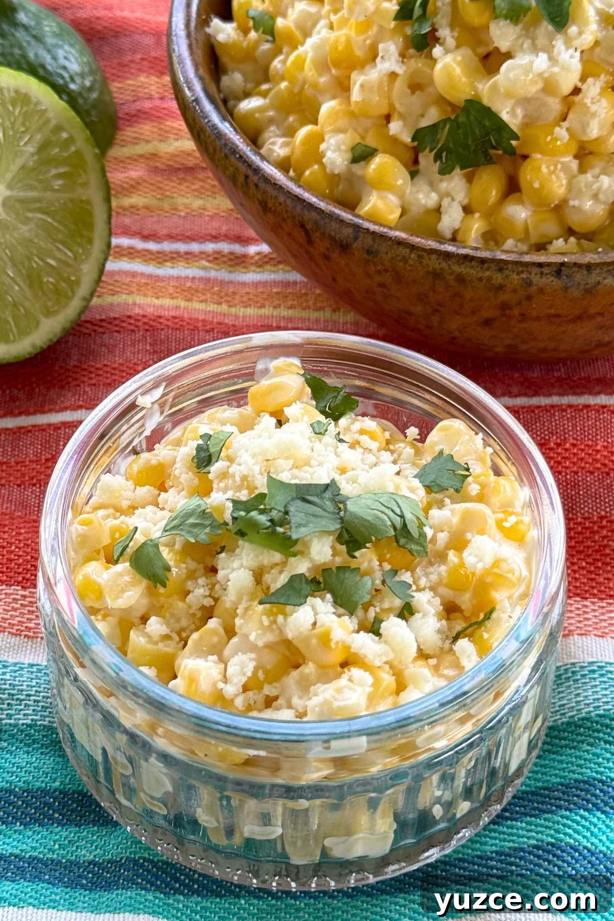 Mexican street corn in a glass cup with a lime and a bowl with more street corn behind it.