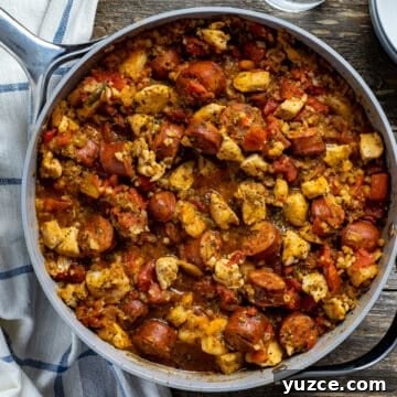 cooked jambalaya in pan on wooden table with glass of water and white bowl on the side and blue and white striped kitchen towel in the background
