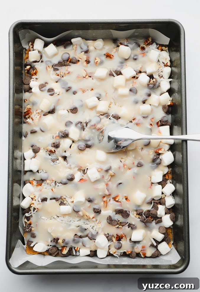 a spoon spreading condensed milk over layers of candy and chocolate in a metal baking pan.