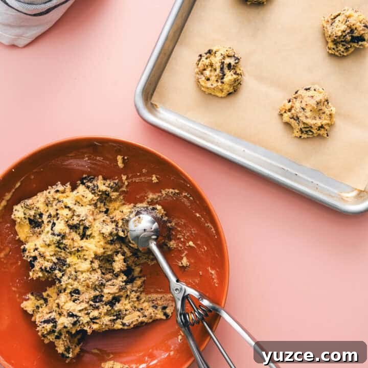 Chocolate Almond Cookie Recipe with Crunchy Almonds and Cocoa 6 Cookie dough with chopped chocolate and almonds in a bowl, being portioned with a scoop onto a baking tray lined with parchment paper.