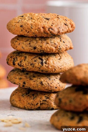 Stack of chocolate almond cookies showing their golden texture and chocolate flecks, with more cookies blurred in the background on a soft pink surface.