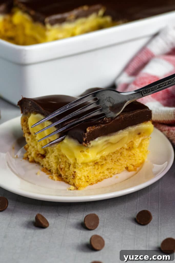 Boston Cream Pie Cake from a Box Mix — Quick Layered Dessert 5 A fork cutting into a piece of Boston cream pie cake on a white plate.