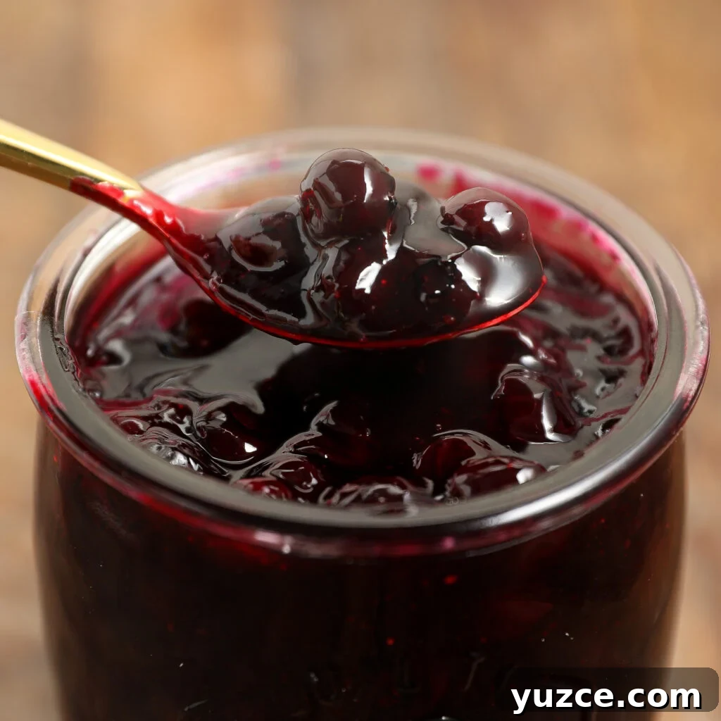 Homemade Blueberry Pie Filling Recipe for Perfect Pies 2 A close-up of a glass jar filled with thick, glossy blueberry jam or blueberry pie filling. A spoonful, showing whole berries, is held above the jar. The background is softly blurred brown wood.
