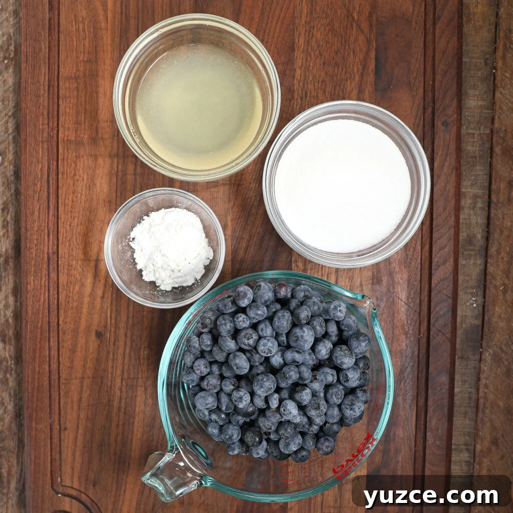 Homemade Blueberry Pie Filling Recipe for Perfect Pies 3 A measuring cup filled with fresh blueberries sits on a wooden surface, ready to become blueberry pie filling, next to small glass bowls containing lemon juice, granulated sugar, and cornstarch.