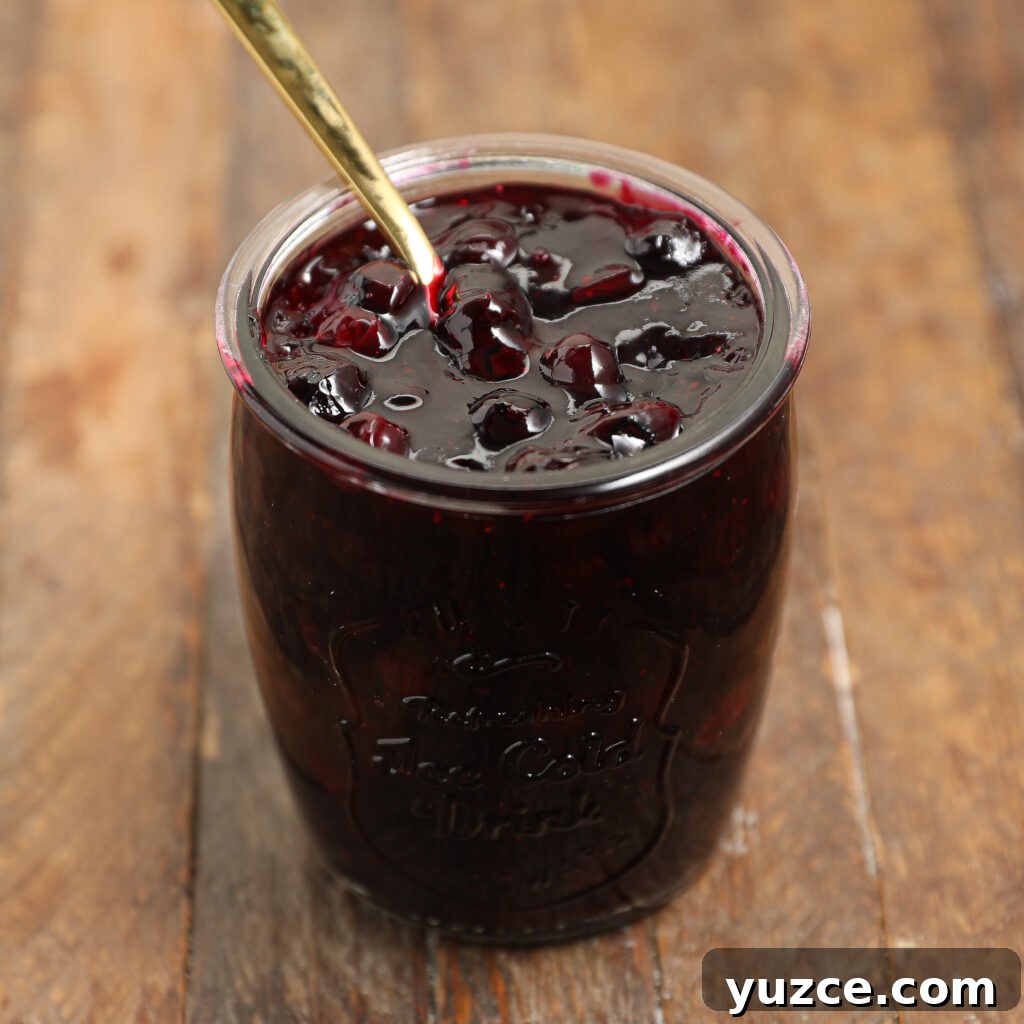 Homemade Blueberry Pie Filling Recipe for Perfect Pies 4 A glass jar filled with dark berry jam, reminiscent of blueberry pie filling, sits on a wooden surface. A gold spoon is resting in the jam, which contains visible whole berries.