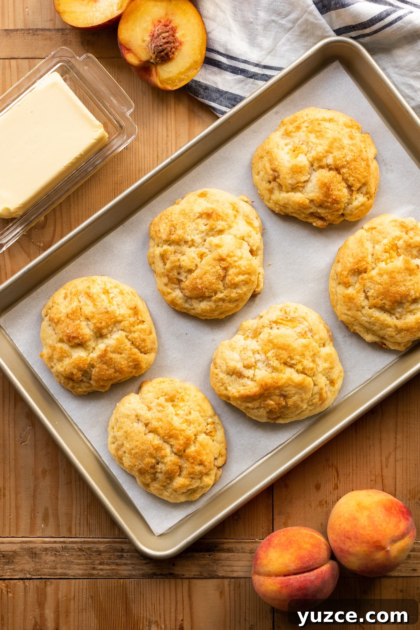 Top down view of peach drop biscuits cooling on piece of parchment paper in a baking tin with butter and sliced peaches in background all on wooden surface