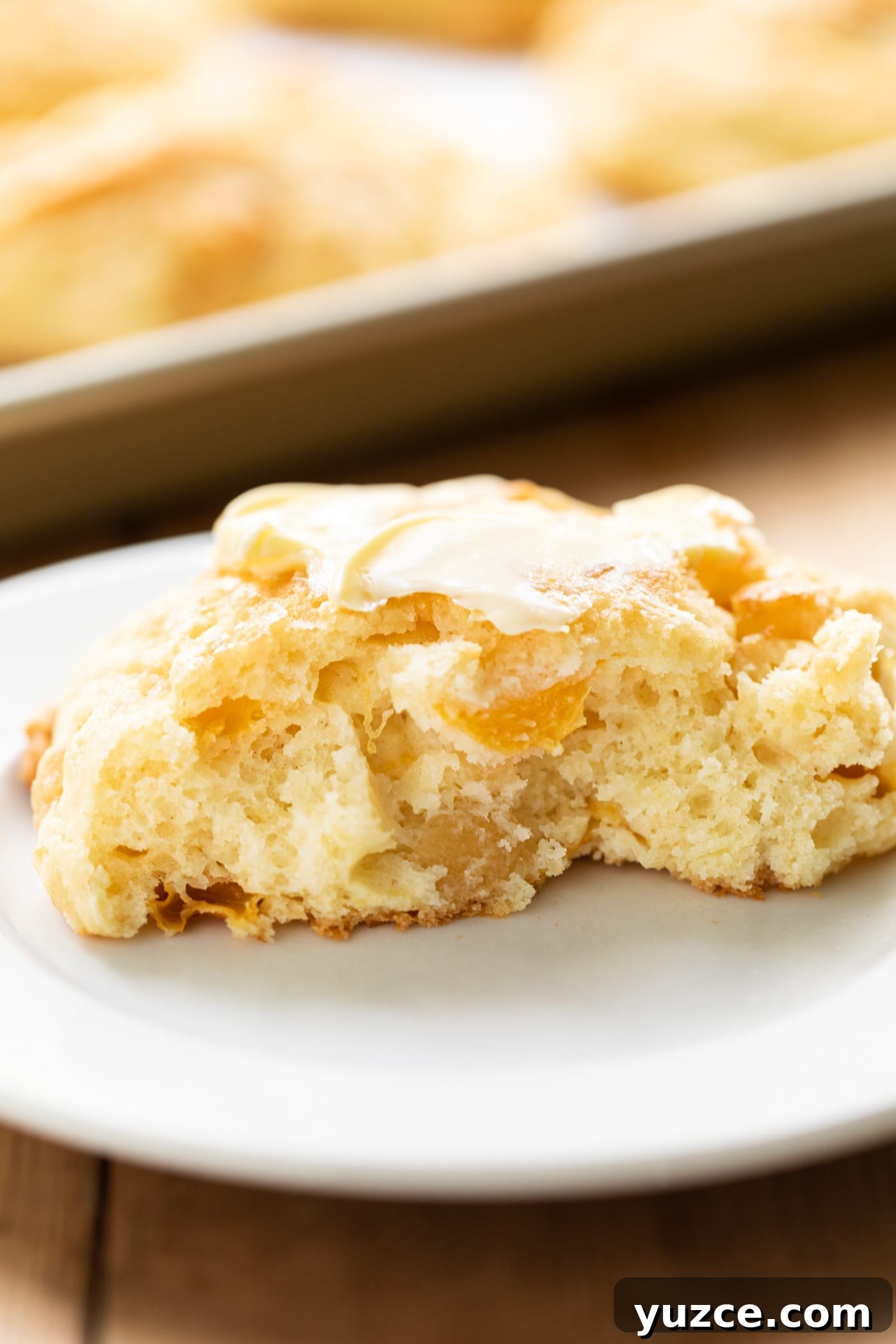Peach drop biscuit cut in half with center exposed slathered with butter on white plate with extra biscuits on baking sheet in background all on wooden surface