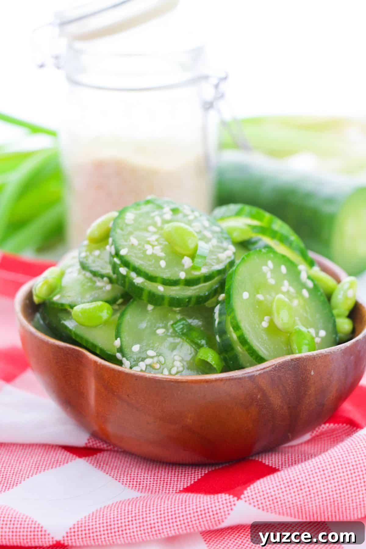 Cucumber and Edamame Salad with Sesame Ginger Dressing 2 A wooden bowl filled with Cucumber Edamame Salad, topped with sesame seeds and set on a red and white checkered cloth. A jar and cucumber rest in the background.