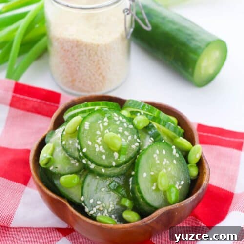 Cucumber and Edamame Salad with Sesame Ginger Dressing 11 A wooden bowl of Cucumber Edamame Salad topped with sesame seeds sits on a red and white checkered cloth, with cucumber, green onions, and a jar of sesame seeds in the background.