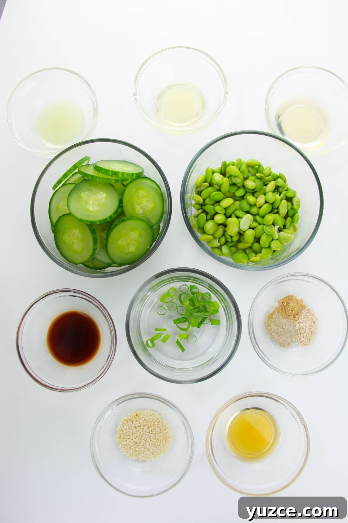 Cucumber and Edamame Salad with Sesame Ginger Dressing 4 A top view of glass bowls containing ingredients sliced cucumber, edamame, chopped green onions, and various sauces, seasonings, and liquids arranged on a white surface.