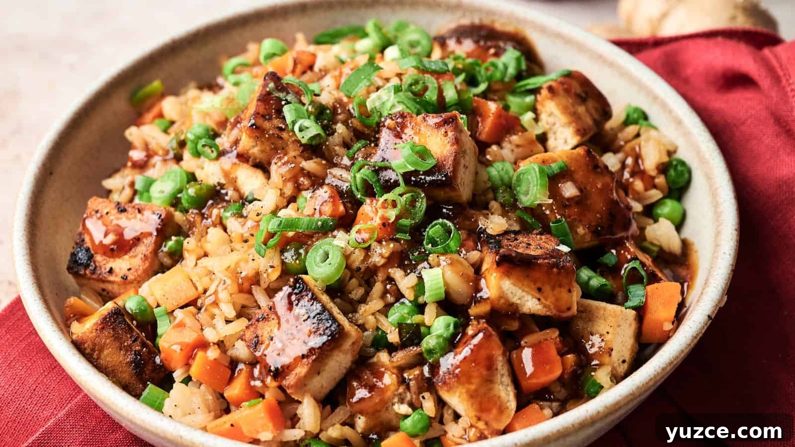 A bowl of fried rice with grilled tofu, diced carrots, peas, chopped green onions, and a drizzle of dark sauce, served on a red cloth.