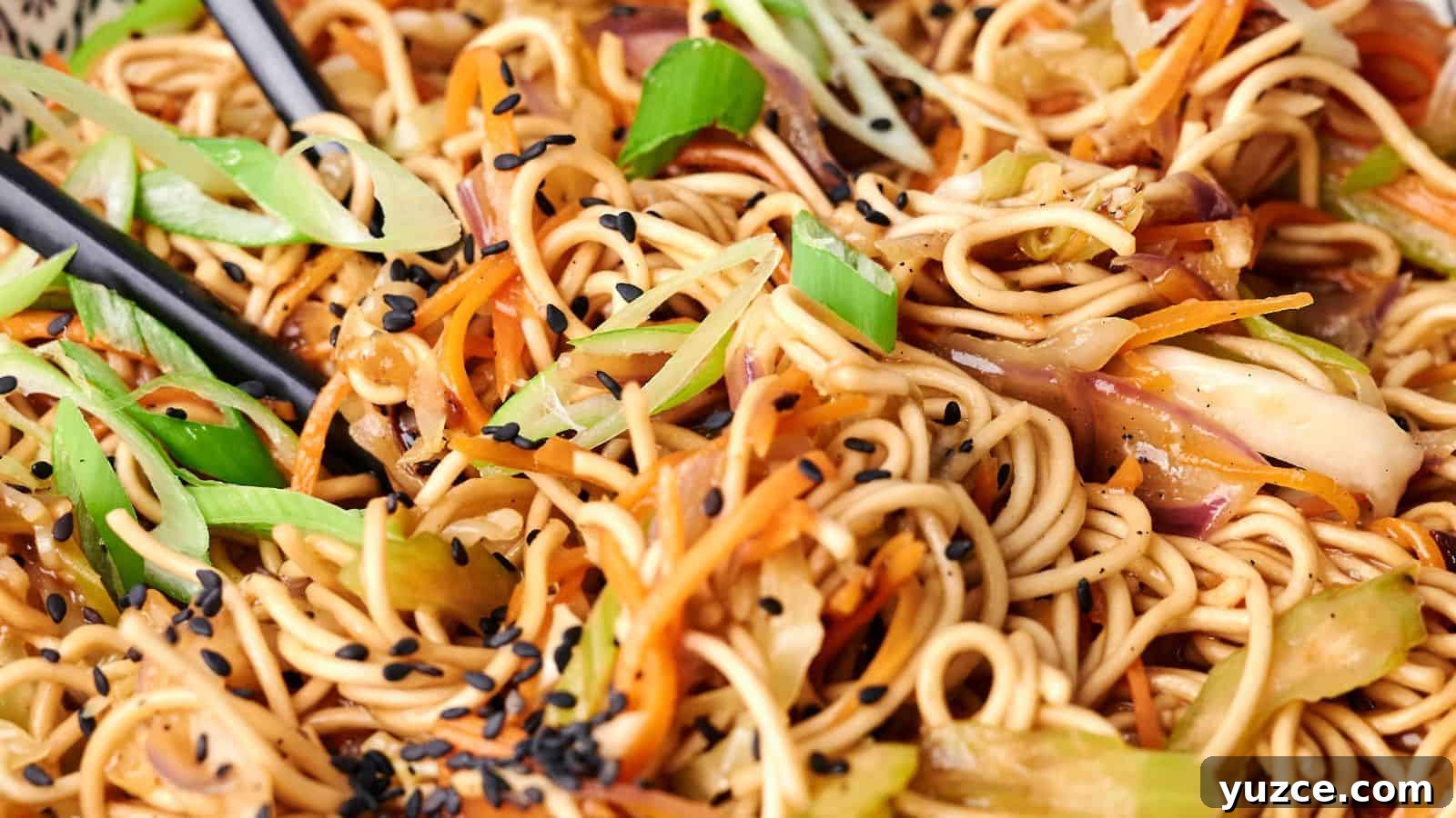 A close-up view of a plate of chow mein stir-fried noodles with vegetables and black sesame seeds, with chopsticks visibly picking up a portion.
