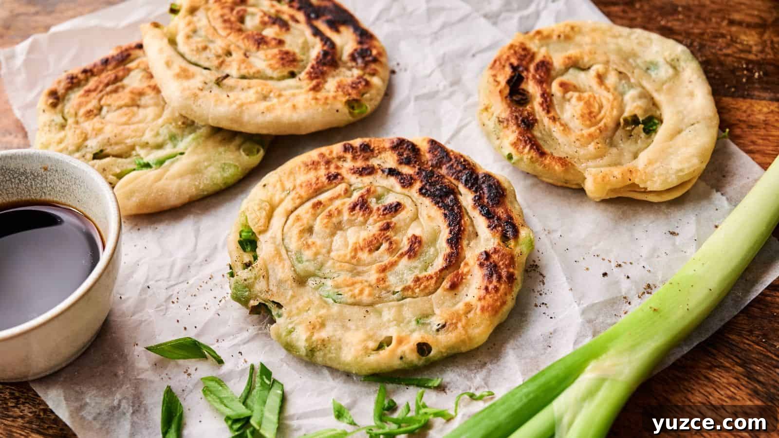 Four cooked scallion pancakes on parchment paper, garnished with sliced green onions, with a small bowl of dipping sauce and a whole scallion beside them.