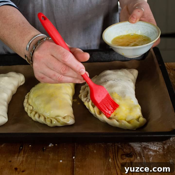 Authentic Cornish Pasty Recipe: Traditional Step-by-Step Guide 14 womans hands brushing beaten eggs with a red silicone brush onto three uncooked Cornish pastys on a black baking tray