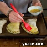Authentic Cornish Pasty Recipe: Traditional Step-by-Step Guide 23 womans hands brushing beaten eggs with a red silicone brush onto three uncooked Cornish pastys on a black baking tray