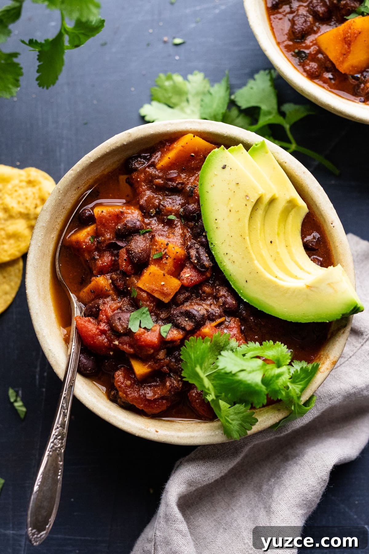 Hearty Sweet Potato and Black Bean Chili Recipe 2 A ceramic bowl with sweet potato black bean chili in it, topped with avocado slices and cilantro leaves. The bowl is arranged on a gray background with tortilla chips, a gray napkin, and cilantro placed around the bowl.