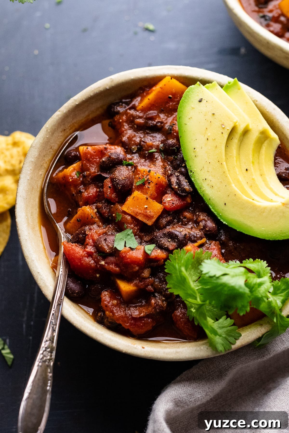 Hearty Sweet Potato and Black Bean Chili Recipe 8 A ceramic bowl with sweet potato black bean chili in it, topped with avocado slices and cilantro leaves. The bowl is arranged on a gray background with tortilla chips, a gray napkin, and cilantro placed around the bowl.