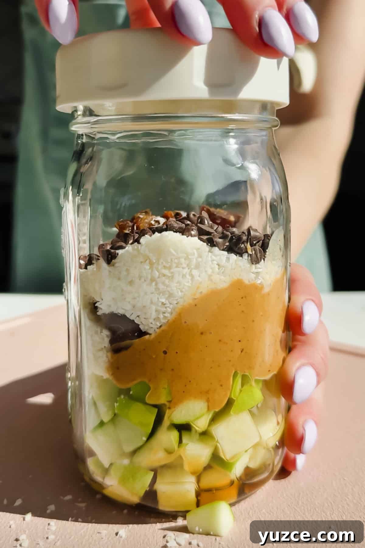 Caramel deLites Apple Salad in a Jar — Layered Dessert Salad Recipe 3 A hand holds a glass jar filled with chopped green apple, peanut butter, shredded coconut, chocolate chips, and cacao nibs, viewed from the side, with a lid being placed on top.