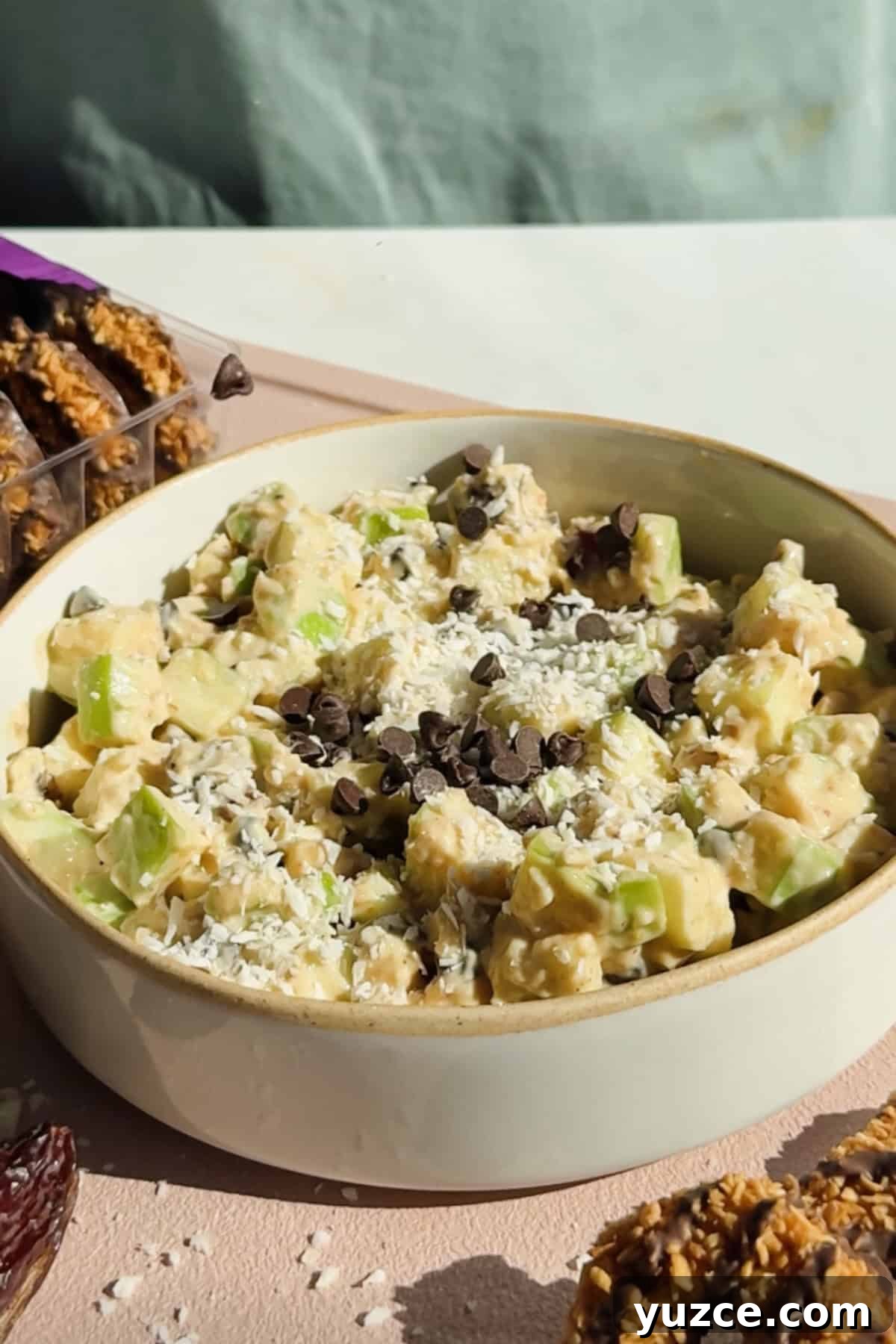 Caramel deLites Apple Salad in a Jar — Layered Dessert Salad Recipe 4 A bowl of apple salad with diced apples, shredded coconut, and chocolate chips on top, surrounded by granola bars and cookies.