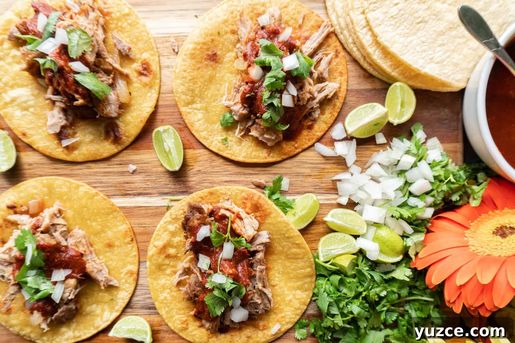 Authentic Pork Carnitas Street Tacos Recipe 10 four carnitas tacos topped with red chile sauce, chopped onion, and cilantro, on a wooden cutting board.