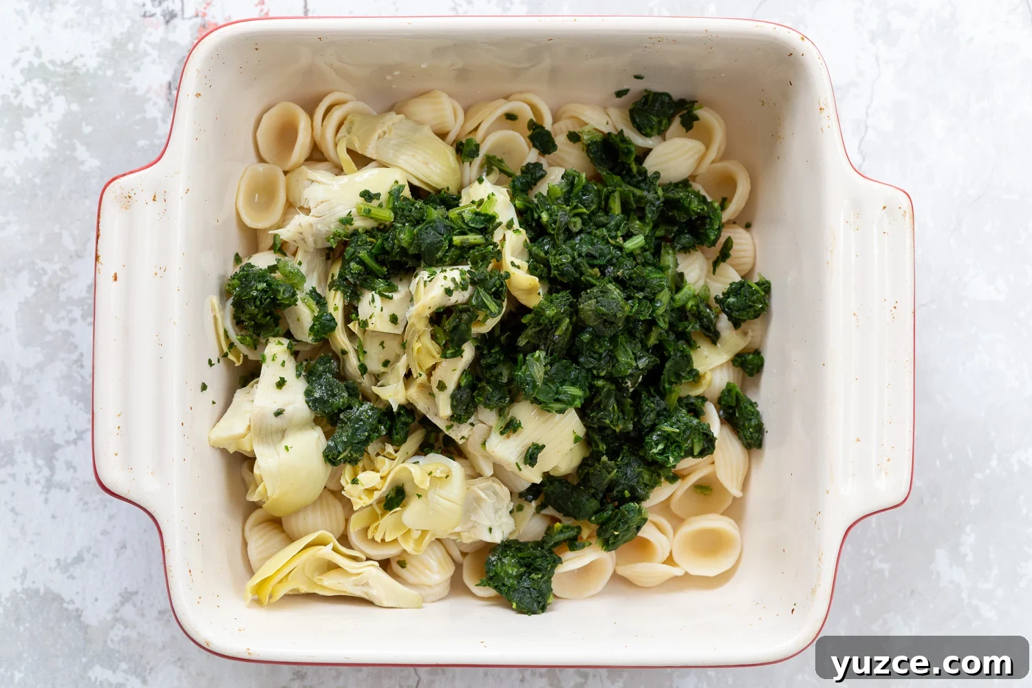 spinach pasta recipe overhead shot of a casserole dish with cooked shell pasta, spinach, and chopped artichokes
