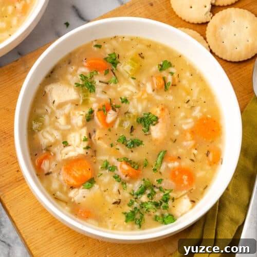 A bowl of turkey and rice soup sitting on a wood board next to some crackers.