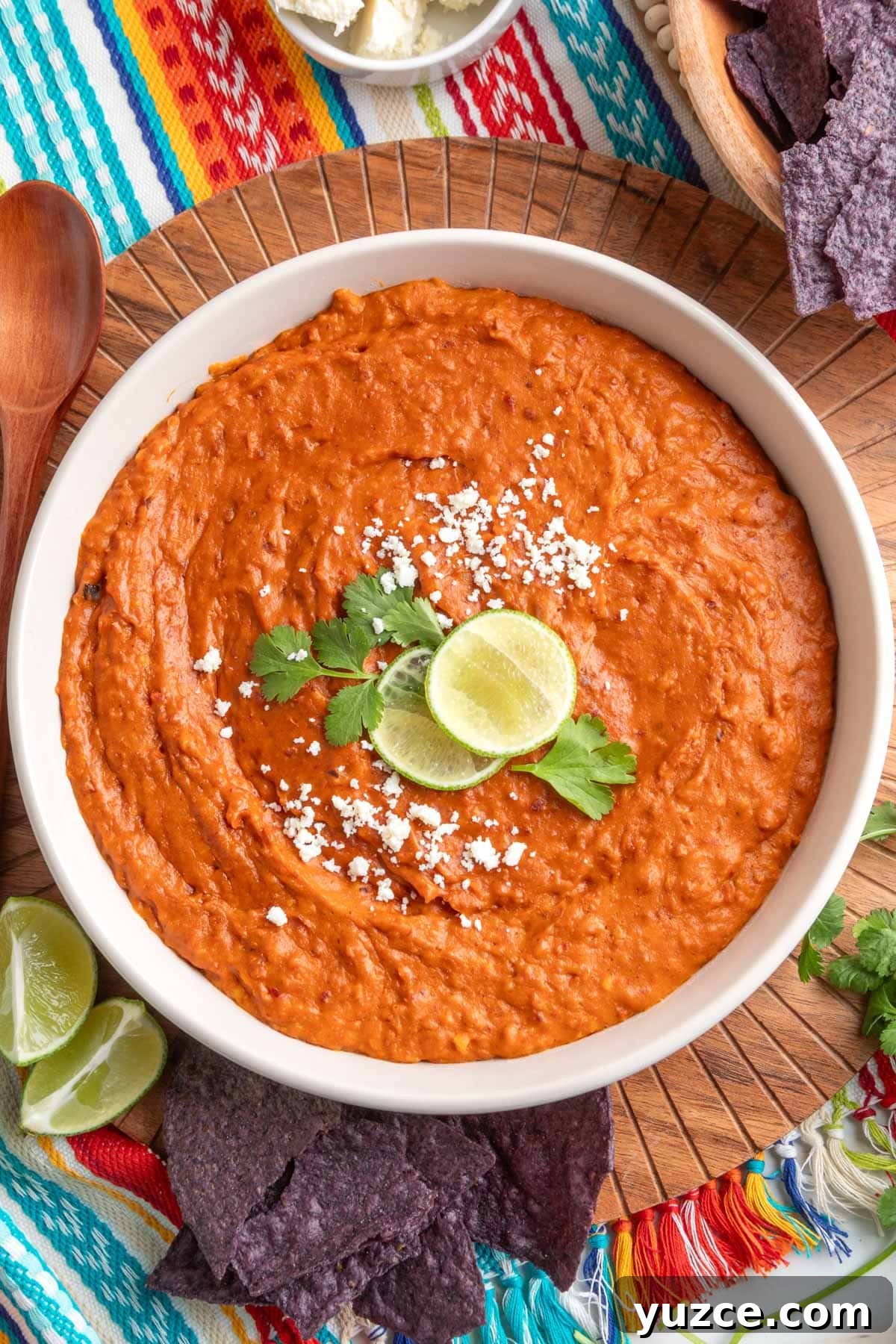 Chipotle Chorizo Bean Dip Recipe — Smoky Spicy Party Dip 2 Overhead view of creamy chorizo bean dip in a white bowl, topped with crumbled cheese, lime slices, and cilantro, surrounded by blue corn tortilla chips on a colorful striped cloth.