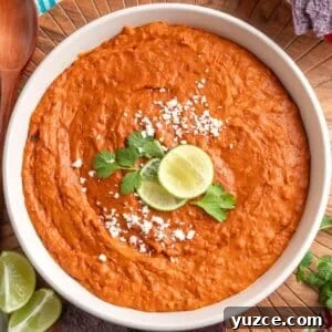 Chipotle Chorizo Bean Dip Recipe — Smoky Spicy Party Dip 10 Creamy chorizo bean dip in a white bowl, garnished with crumbled cheese, cilantro, and lime slices, with blue corn tortilla chips and a wooden spoon on the side.