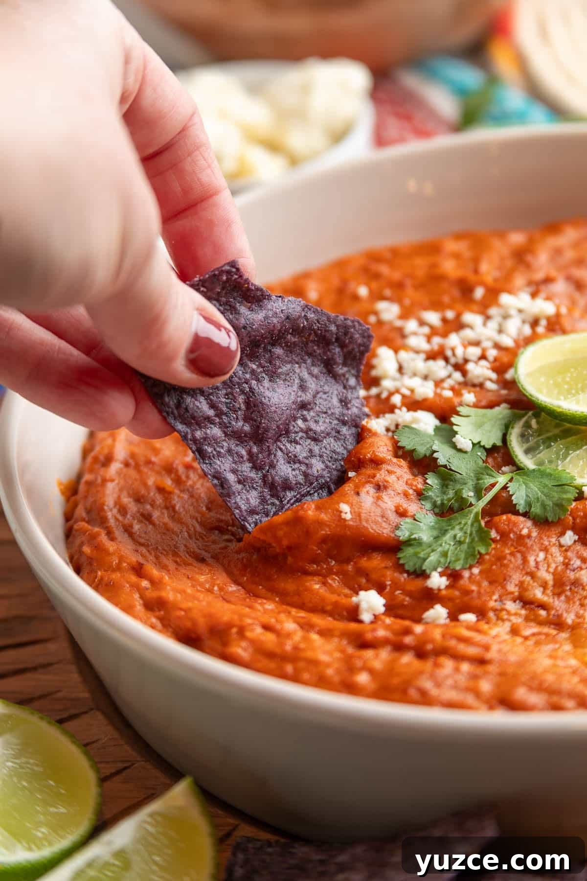 Chipotle Chorizo Bean Dip Recipe — Smoky Spicy Party Dip 8 Hand dipping a blue corn tortilla chip into creamy chorizo bean dip topped with crumbled cheese, cilantro, and lime slices.