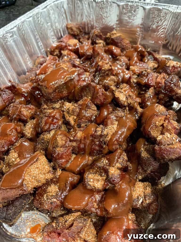 Smoky Pork Shoulder Burnt Ends Recipe for Tender, Crispy Bites 5 pork shoulder chunks in an aluminum pan with rub,, barbecue sauce, and honey