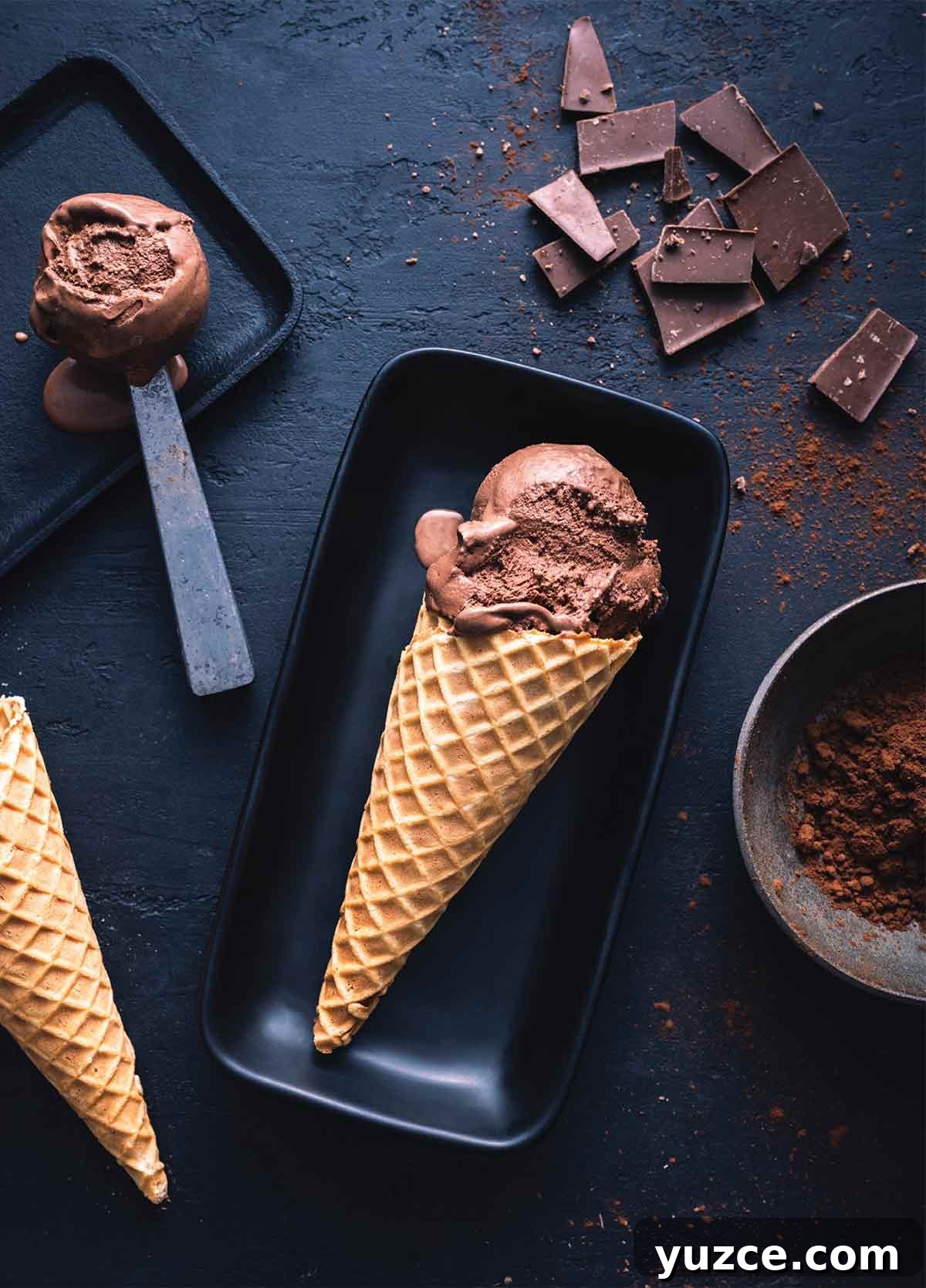 Decadent Dark Chocolate Gelato Recipe — Creamy Italian Dessert 2 2 rectangular plates with scoops of dark chocolate gelato, one on a scoop and one in a cone, surrounded by squares of dark chocolate and a bowl of cocoa.