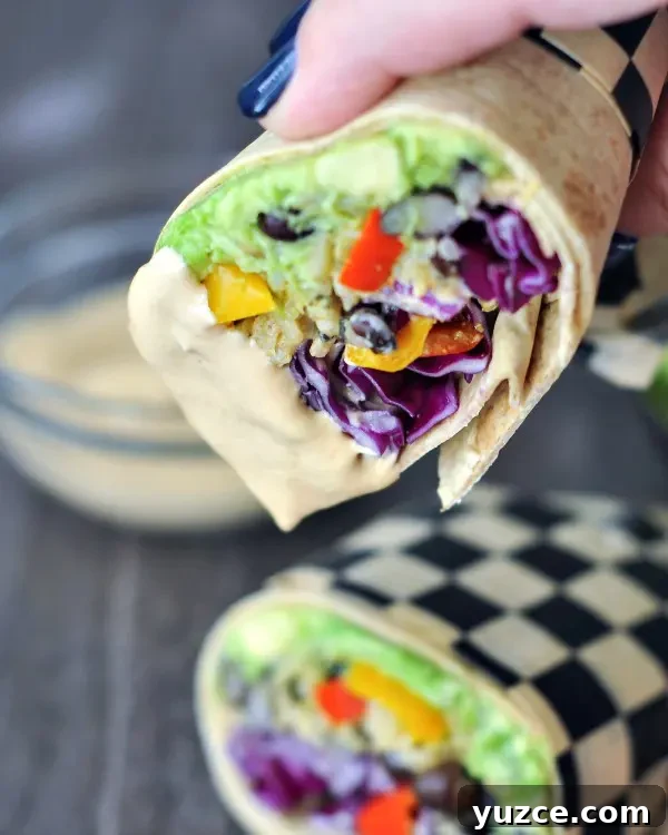 Colorful vegan burrito sliced in half and dipped into spicy chipotle sauce, half of the burrito held in a hand, other half wrapped in checkered paper and sitting on a dish.