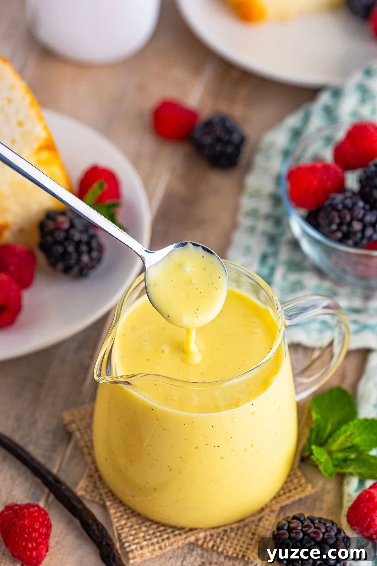 A small silver spoon drizzling Creme Anglaise Recipe back into the small glass with pour spout. Sitting on wooden table top, surround by berries and mint.